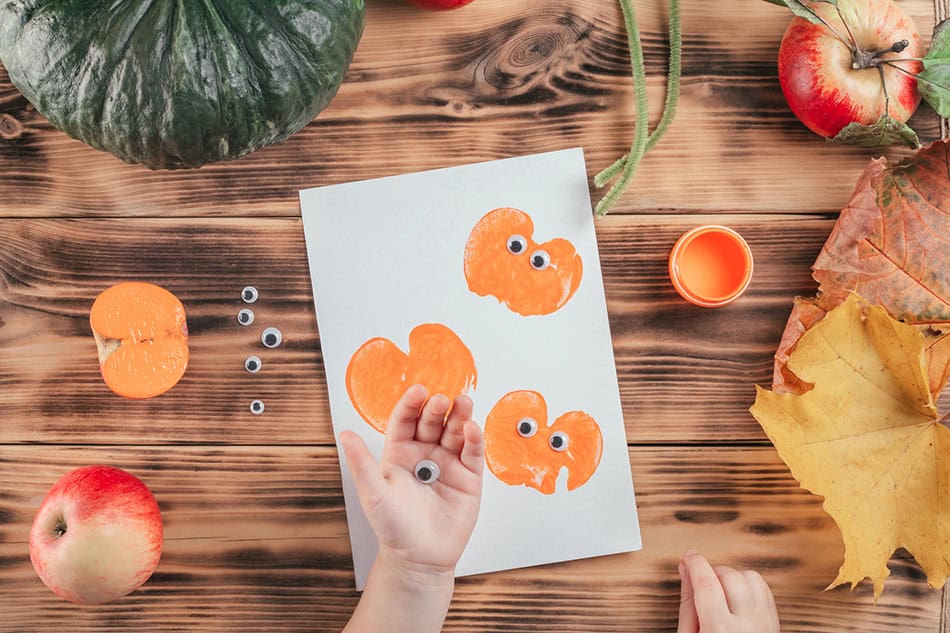 Step-by-step Halloween childrens tutorial pumpkin apple prints. Step 11: Child hand holds eyes to glue to pumpkin print